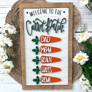 May include: Welcome to the Carrot Patch sign with a wooden frame and white background. The sign has five carrot-shaped pieces with green tops and names on each: Dad, Mom, Beaux, Hayes, and Remi.