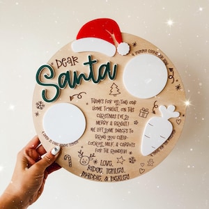 May include: A wooden Christmas Eve plate with the words "Dear Santa" in green and white. It includes spaces for cookies, milk, and carrots, along with a Santa hat and festive decorations. The plate is held by a person.