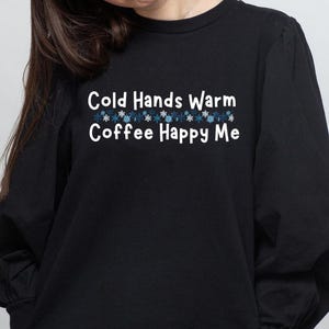 以下が含まれることがあります： 「Cold Hands Warm Coffee Happy Me」の白文字がプリントされた黒の長袖スウェットシャツ。テキストの上には青と白の雪の結晶のデザインが並んでいます。スウェットシャツは丸首で、袖はパフスリーブです。