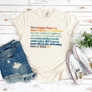 Op de afbeelding: Een beige T-shirt met een regenboogkleurige afbeelding met de tekst "Ik ben een happy flaps toy lines fidgets co-regulation no eye contact required alternate communication accommodating needs embracing differences neurodiversity affirming kind of BCBA ∞".
