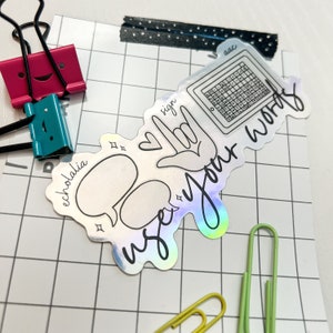 Puede incluir: Una pegatina holográfica con las palabras "use your words" en una fuente manuscrita. La pegatina incluye bocadillos, un teclado y un signo de mano. Otros elementos en la imagen incluyen clips y clips de papel.