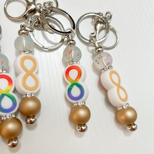 May include: Four keychains with rainbow infinity symbol charms, gold beads, and clear beads with iridescent glitter. Each keychain has a silver split ring.