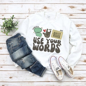 Puede incluir: Sudadera blanca con el texto "USE YOUR WORDS" en letras marrones. El texto está rodeado de ilustraciones de burbujas de diálogo, una mano firmando y un dispositivo de comunicación.