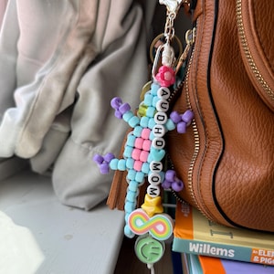 May include: A beaded lizard keyring featuring light blue and pink plastic beads, with white letter beads spelling AUDHD MOM. The charm includes a pink flower, a yellow star, a rainbow infinity symbol, and a green smiley face bead on a metal clip.