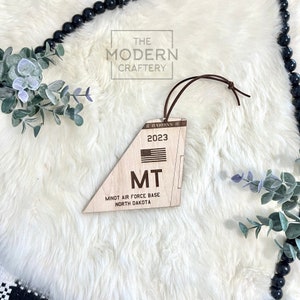 May include: Wooden ornament shaped like a plane wing with the text "MT MINOT AIR FORCE BASE NORTH DAKOTA" and "2023" with a small American flag.