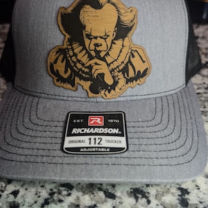 May include: Gray and black trucker hat with a wooden-style patch featuring a detailed image of a clown. The hat has a mesh back and an adjustable strap. The patch has a detailed image of a clown. The hat is labeled "RICHARDSON 112 TRUCKER".