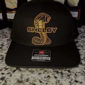 May include: Black trucker hat with a gold cobra logo and the name "SHELBY". The hat has a mesh back and a curved brim. The Richardson brand tag is visible on the front.