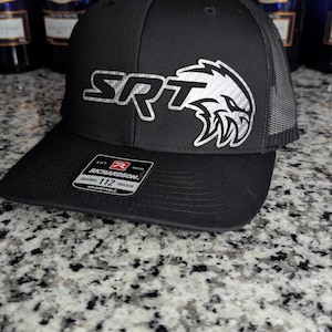 May include: Black Richardson 112 trucker hat with the letters "SRT" and a silver eagle logo on the front. The hat has a mesh back and a curved brim. The hat is sitting on a speckled surface.