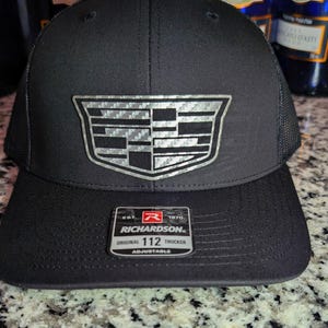May include: Black trucker hat with a silver emblem. The hat features a curved brim and a mesh back. The emblem is a silver Cadillac logo. The hat has a Richardson tag that says "Original 112 Trucker Adjustable".