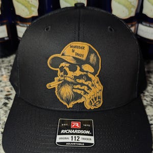 May include: Black Richardson 112 trucker hat with a skull graphic wearing a hat and smoking a cigar. The graphic includes the text "WHATEVER IT TAKES". Adjustable hat.