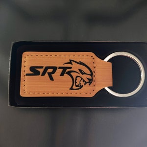 May include: A brown leather keychain with a black SRT logo and a silver key ring. The keychain is in a black box.