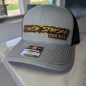May include: Gray and black trucker hat with the words "POWER STROKE DIESEL" in gold and black lettering. The hat has a curved brim and a mesh back. The hat is adjustable and has a Richardson tag.