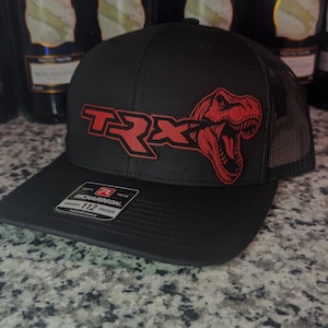 May include: Black trucker hat with a red "TRX" logo and a red dinosaur graphic. The hat has a mesh back and a curved brim. The Richardson 112 Trucker hat is adjustable.