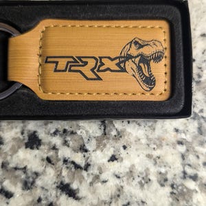 May include: A tan leather keychain with the word "TRX" and a Tyrannosaurus Rex graphic in black. The keychain is in a black box, with a silver key ring attached.