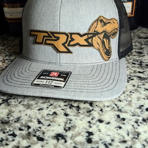 May include: Gray and black trucker hat with a brown and gold TRX logo featuring a dinosaur head. The hat has a curved brim and a mesh back. The Richardson 112 Trucker hat is adjustable.