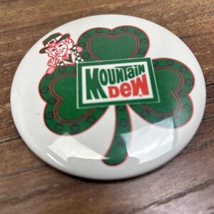May include: A round, white Mountain Dew button featuring a green shamrock design with a leprechaun and the Mountain Dew logo. The shamrock has a red outline and the text is in green and red.