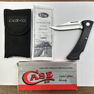 May include: A black folding knife with a silver blade, a black fabric sheath with the text CASE XX, a white paper insert labeled CASE HAND-CRAFTED KNIVES SINCE 1889, and a rectangular box with the text CASE XX and CASE. FIRST CHOICE OF KNIFE COLLECTORS.