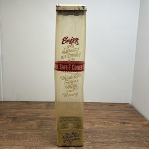 May include: A vintage ice cream cone dispenser with text reading "Enjoy Our Quality Ice Cream With Safe-T Cones." The dispenser is rectangular, with a clear plastic front and a metal frame. The bottom reads "This Store Sells the Best of Everything."