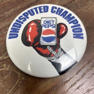 May include: A white button with the words "UNDISPUTED CHAMPION" in blue. The button features a red and black boxing glove holding a Diet Pepsi can. The can has a red, white, and blue design.