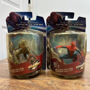 Op de afbeelding: Twee The Amazing Spider-Man deluxe actiefiguren in originele verpakking. De ene blisterverpakking bevat een groene hagedisfiguur en de andere een rood-blauwe Spider-Man-figuur. Beide verpakkingen zijn voorzien van karakterafbeeldingen en logo's.