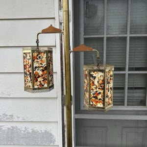 Könnte beinhalten: Zwei Vintage-Hängelampen mit einem Blumenmuster in Orange, Schwarz und Weiß. Die Lampen haben einen Messingrahmen und eine dekorative Metallverkleidung. Die Lampen sind an einer Messingstange mit Holzakzenten befestigt.