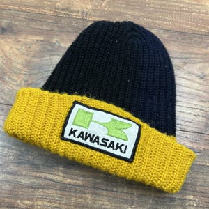 Może przedstawiać: Czarna i żółta dzianinowa czapka z naszywką z logo Kawasaki. Czapka ma prążkowany żółty brzeg i czarną koronę. Logo Kawasaki jest zielono-białe.