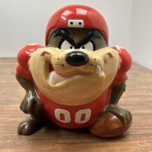May include: A ceramic Tazmanian Devil figure in a football uniform. The figure is wearing a red helmet and jersey with the number "00". The football is brown, and the figure has an angry expression.