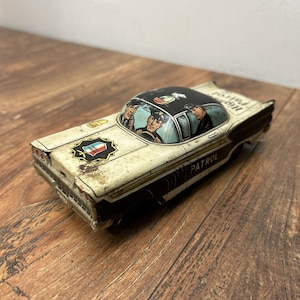 Pode incluir: Carro de polícia de brinquedo vintage em creme, preto e branco. O carro apresenta uma ilustração detalhada de oficiais no interior e a palavra "PATROL" na lateral. O teto é preto com uma cena pintada. O carro tem um distintivo na frente.