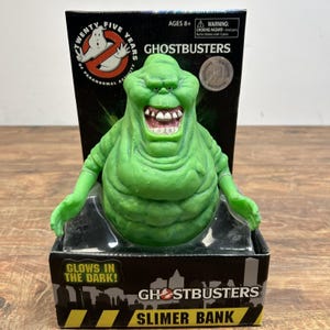Peut inclure: Une tirelire verte représentant le personnage Slimer de Ghostbusters dans son emballage noir d'origine. La figurine a un large sourire édenté et une peau texturée. La boîte affiche le logo Ghostbusters, le texte Twenty Five Years of Paranormal Activity et Glows in the Dark.