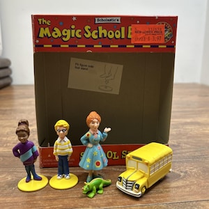 Könnte beinhalten: Ein Magic School Bus Spielzeugset mit einem roten Pappkarton, drei Kunststofffiguren auf gelben runden Sockeln, einer kleinen grünen Echsenfigur und einem gelben Schulbus-Spielzeug. Auf der Schachtel ist der Text The Magic School Bus zu sehen.
