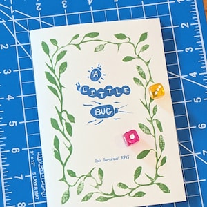 May include: A white booklet with a green leafy border and the title "A Little Bug" in blue lettering. The booklet is on a blue cutting mat with a ruler and two dice, one yellow and one pink.