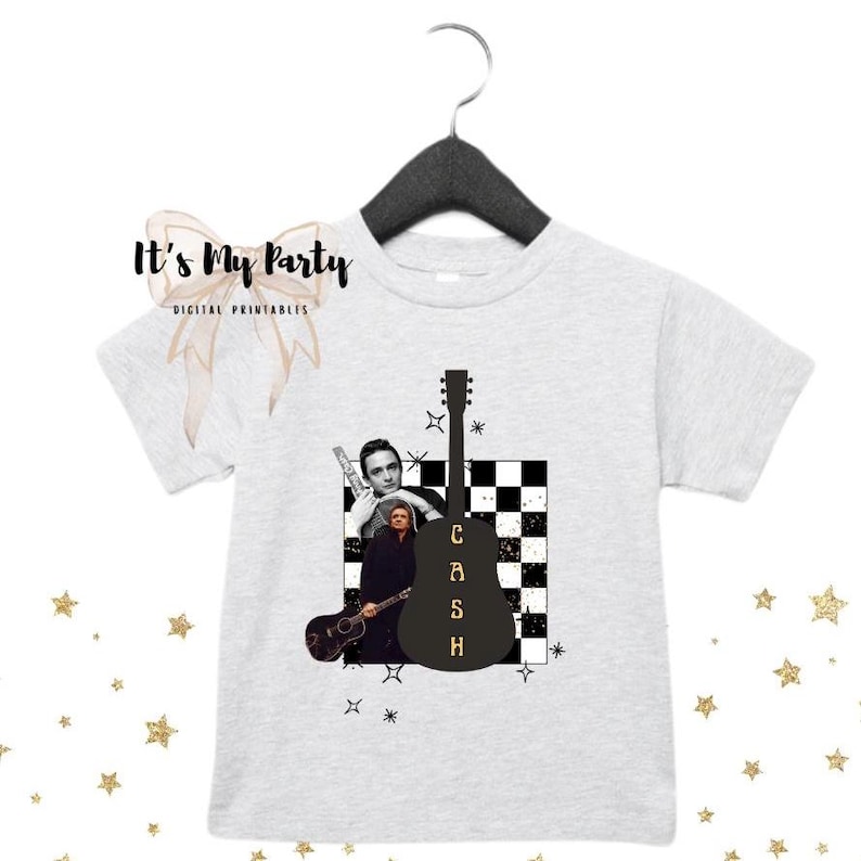 May include: A light gray t-shirt featuring a graphic of a black guitar with the word "CASH" in gold letters. The shirt also includes images of a musician and a checkered background. The shirt is hanging on a black hanger.