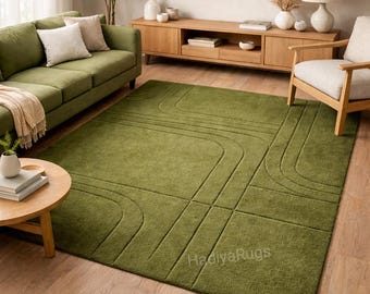 May include: A large, olive green area rug with a geometric pattern of curved and straight lines. The rug is placed on a hardwood floor in a living room setting, alongside a green sofa and wooden furniture.