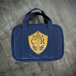 May include: A navy blue carrying case with a wooden shield design. The shield features a sword and the letters "CR" in a gold color. The case has a handle and is rectangular in shape.