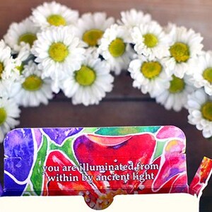 Puede incluir: Un arreglo floral con una corona de margaritas blancas con centros amarillos. Una caja colorida con el texto "you are illuminated from within by ancient light" est&aacute; en primer plano. La imagen tiene una est&eacute;tica natural y brillante.