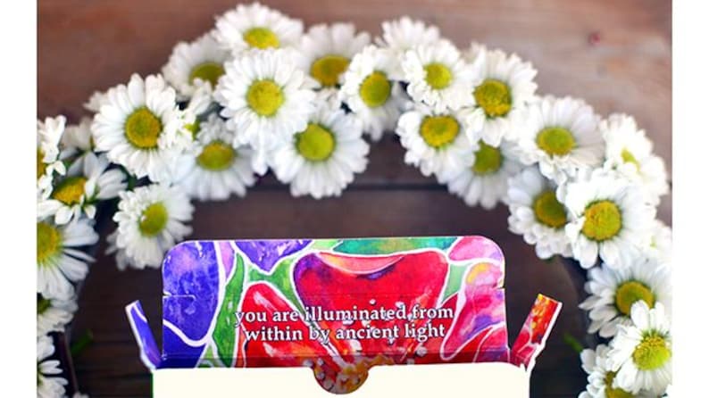 Puede incluir: Un arreglo floral con una corona de margaritas blancas con centros amarillos. Una caja colorida con el texto "you are illuminated from within by ancient light" est&aacute; en primer plano. La imagen tiene una est&eacute;tica natural y brillante.