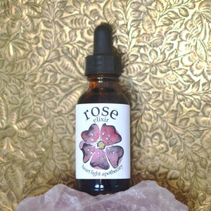May include: A small brown glass bottle with a dropper top. The label is white with the words "rose elixir" and "heart light apothecary" in black lettering. A pink rose with a watercolour effect is in the centre of the label.