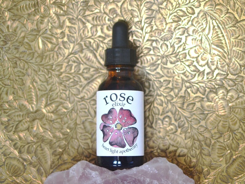 May include: A small brown glass bottle with a dropper top. The label is white with the words "rose elixir" and "heart light apothecary" in black lettering. A pink rose with a watercolour effect is in the centre of the label.