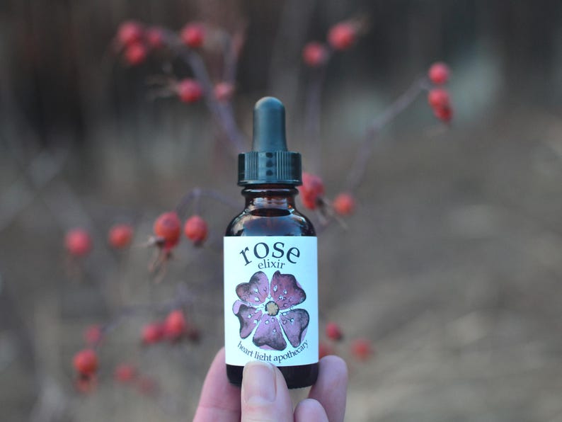 May include: A small brown glass bottle with a dropper top and a white label that says "rose elixir" and "heart light apothecary". The label also features a pink rose with water droplets.
