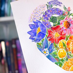 Puede incluir: Pintura a la acuarela de una forma de coraz&oacute;n llena de flores coloridas. La obra presenta flores azules, rojas, naranjas y amarillas con follaje verde. El fondo est&aacute; borroso, sugiriendo una estanter&iacute;a.