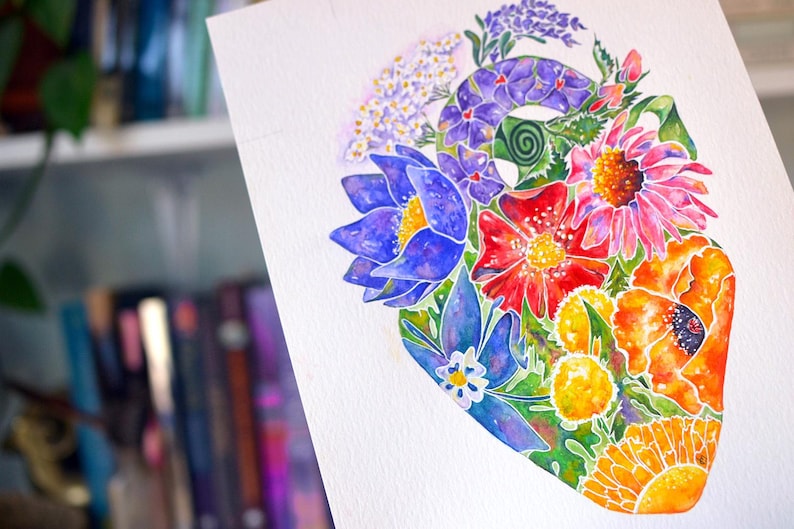 Puede incluir: Pintura a la acuarela de una forma de coraz&oacute;n llena de flores coloridas. La obra presenta flores azules, rojas, naranjas y amarillas con follaje verde. El fondo est&aacute; borroso, sugiriendo una estanter&iacute;a.