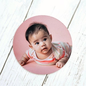 May include: A round pink fabric coaster with a photo of a baby wearing a striped shirt. The baby is looking up at the camera with a curious expression.