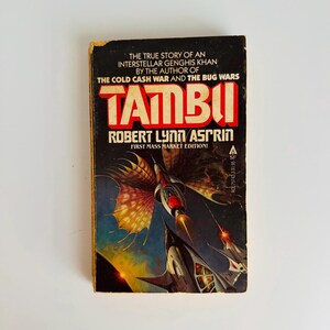 Pu&ograve; includere: Un libro tascabile vintage intitolato "Tambu" di Robert Lynn Asprin. La copertina presenta un'illustrazione di astronave nei toni del blu, nero e rosso. &Egrave; una prima edizione.