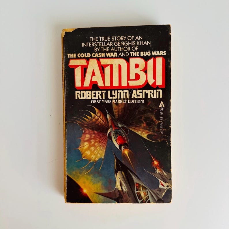 Pu&ograve; includere: Un libro tascabile vintage intitolato "Tambu" di Robert Lynn Asprin. La copertina presenta un'illustrazione di astronave nei toni del blu, nero e rosso. &Egrave; una prima edizione.
