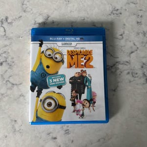 May include: A blue Blu-ray case for "Despicable Me 2" featuring Minions and characters from the animated film. The cover includes the text "BLU-RAY + DIGITAL HD" and "INCLUDES 3 NEW MINI-MOVIES".