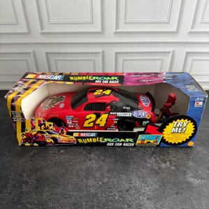May include: A red NASCAR Rumble N Roar Gas Can Racer toy car in its original packaging. The car features the number 24, DuPont branding, and various sponsor logos. The box includes a red gas pump accessory and text reading Try Me.