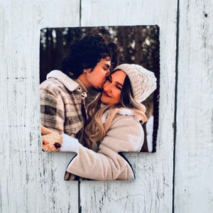 May include: A rectangular can cooler featuring a photograph of a couple embracing and kissing. The man wears a plaid jacket, and the woman wears a white knit hat and a beige coat. The background is a blurred forest scene.