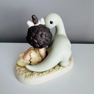 Może przedstawiać: Ceramiczna figurka przedstawiająca dziecko z ciemnobrązowymi włosami i dinozaura. Dziecko ma na sobie jasnoż&oacute;łty str&oacute;j, a dinozaur jest bladozielony. Figurka stoi na kremowej podstawie o fakturze trawy.