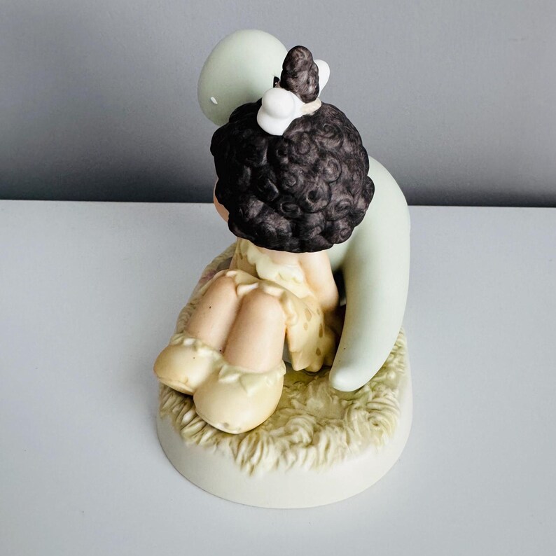 Może przedstawiać: Ceramiczna figurka przedstawiająca dziecko z ciemnymi kręconymi włosami i białą kokardą, obejmujące jasnozieloną formę. Dziecko ma na sobie ż&oacute;łtą sukienkę i siedzi na trawiastym podłożu. Figurka ma matowe wykończenie.