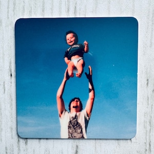 May include: A square coaster featuring a photograph of a person holding a baby up in the air against a bright blue sky. The baby is smiling and wearing a diaper and a dark shirt. The person is wearing a white t-shirt.